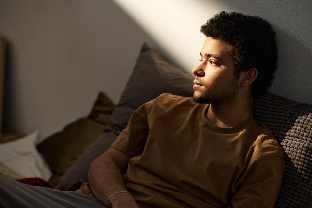 Sad teenage boy worried about his problems sitting alone on bed in his room